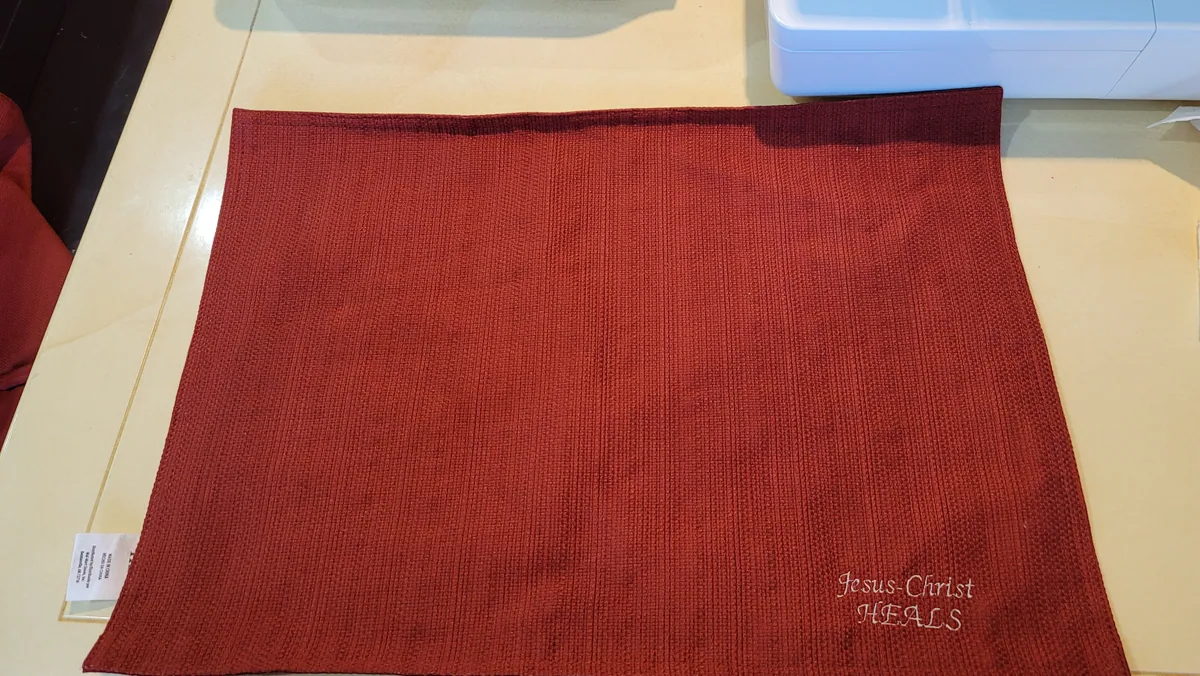 Red waffle-weave placemat with embroidered "Jesus-Christ Heals" in pale thread
